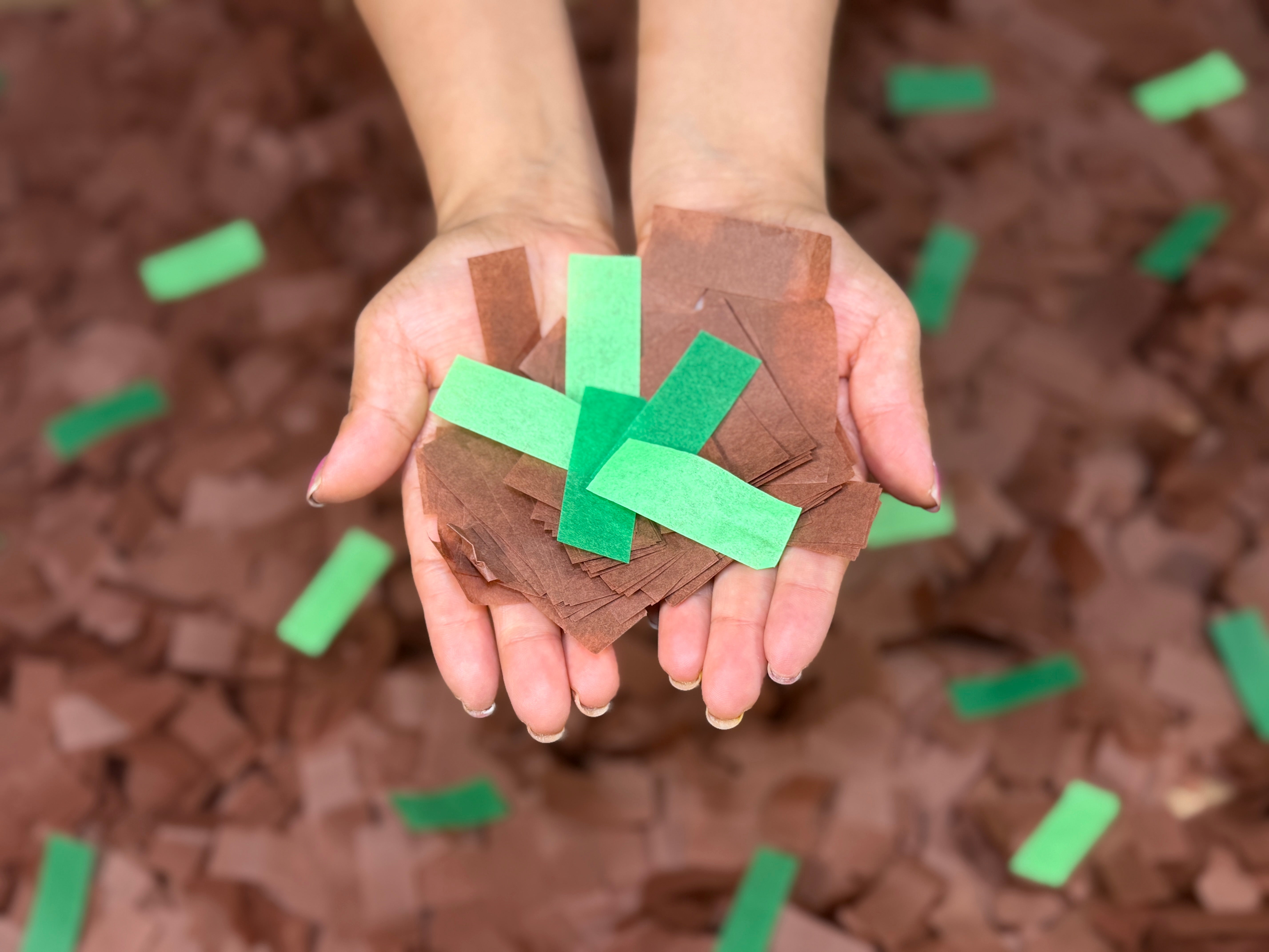 A close-up of a person's hands holding a small tree made from brown paper bark pieces and green paper leaves, with more brown and green pieces scattered in the background.